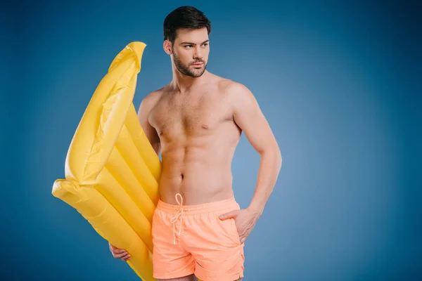 Joven guapo en pantalones cortos sosteniendo colchón de natación y mirando hacia otro lado en azul - foto de stock