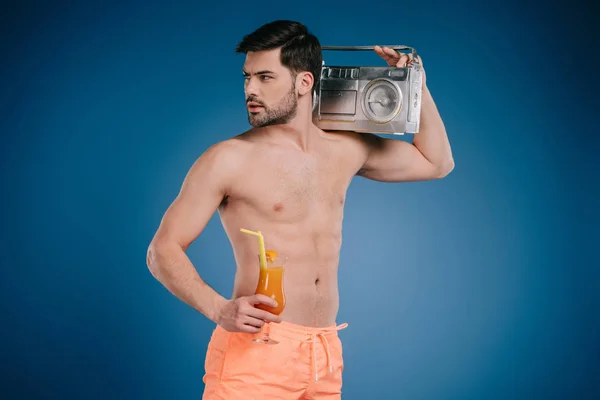 Handsome young man in shorts holding tape recorder and glass of summer cocktail on blue — Stock Photo