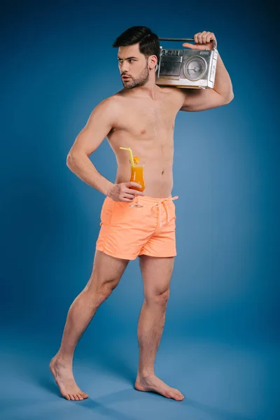 Junger barfüßiger Mann in kurzen Hosen mit Tonbandgerät und einem Glas Sommercocktail auf blauem Grund — Stockfoto