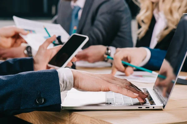 Vista recortada de la mujer de negocios con el teléfono inteligente escribiendo en el ordenador portátil, mientras que los compañeros de trabajo discutir en la mesa en la oficina - foto de stock