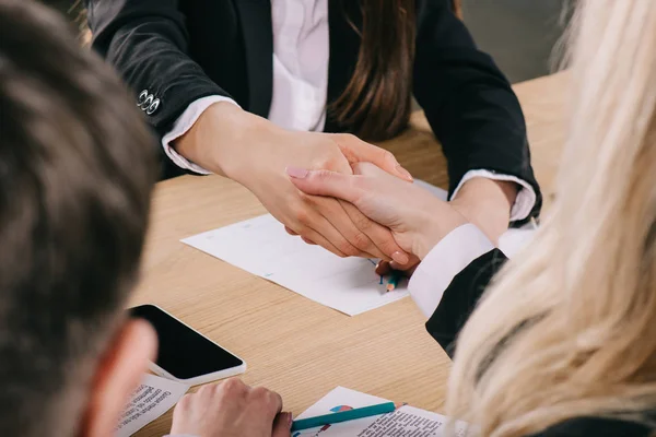 Vista recortada de dos mujeres de negocios estrechando la mano en la mesa en la oficina - foto de stock