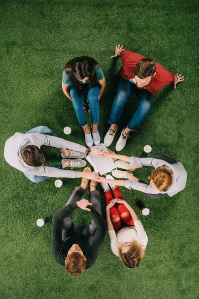 Vista aérea de los socios de negocios tomados de la mano y sentados con otros colegas en la hierba - foto de stock