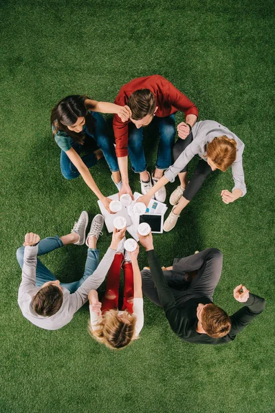 Vue du dessus des partenaires commerciaux cliquetis tasses ensemble tout en étant assis sur l'herbe — Photo de stock