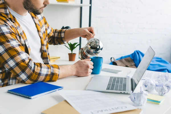 Freelancer verter café en taza en la mesa con el ordenador portátil en la oficina en casa - foto de stock