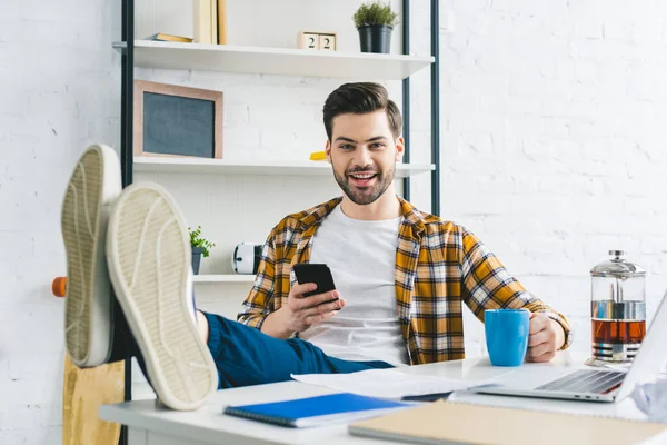 Young freelancer drinking coffee and using smartphone in light office — Stock Photo