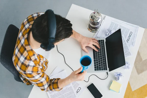 Vista aérea del hombre en auriculares escribiendo en el teclado del ordenador portátil en la oficina de luz - foto de stock