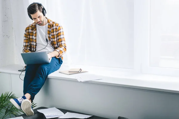 Mann mit Kopfhörer arbeitet am Laptop und sitzt auf Fensterbank — Stockfoto