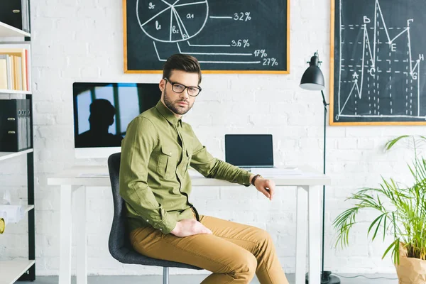 Serious man sitting by table with computers in light office — Stock Photo