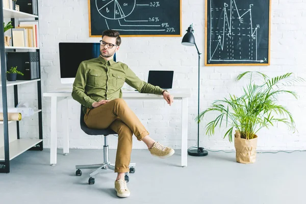 Elegante hombre sentado junto a la mesa con computadoras en la oficina en casa - foto de stock