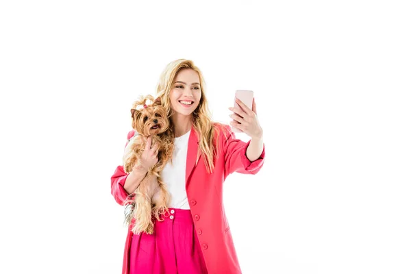 Приваблива молода жінка, одягнена в рожевий, тримає милого собаку і приймає селфі ізольовано на білому — стокове фото