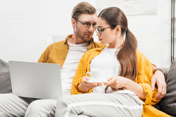 Coppia con caffè e laptop coccolati sul divano di casa — Foto stock