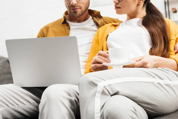 Обрезанный снимок молодой пары с кофе и ноутбуком, сидящих на диване вместе — стоковое фото