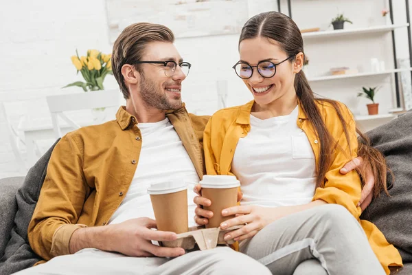 Young happy couple drinking coffee from paper cups on couch — Stock Photo