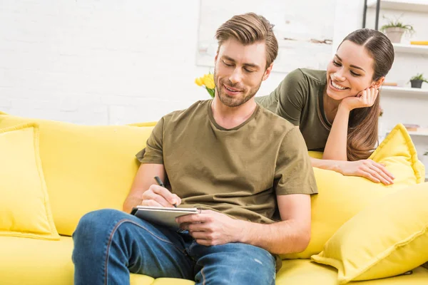 Young man writing in notebook while his girlfriend looking at him from behind on couch at home — Stock Photo