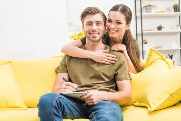 Young man writing in notebook while his girlfriend embracing him from behind on couch at home — Stock Photo