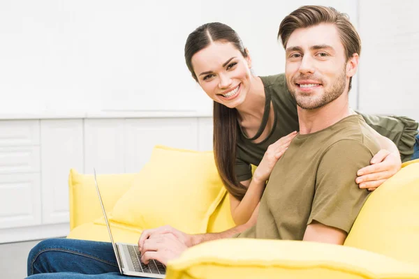 Joven guapo que trabaja en casa con el ordenador portátil, mientras que su novia lo abraza por detrás - foto de stock