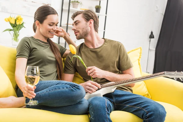 Hombre con guitarra eléctrica presentando flor de tulipán a novia feliz mientras ella sostiene una copa de vino en el sofá en casa - foto de stock