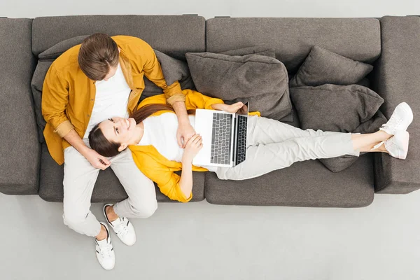 Вид сверху на счастливую женщину лежащую на коленях бойфренда и работающую с ноутбуком на уютном диване дома — стоковое фото