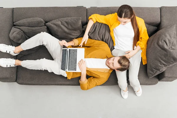 Vista superior del hombre acostado en las rodillas de la novia y el trabajo con el ordenador portátil en el sofá acogedor en casa - foto de stock