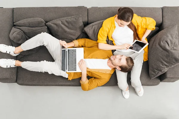 Vue aérienne du couple utilisant des appareils numériques sur un canapé confortable à la maison — Photo de stock