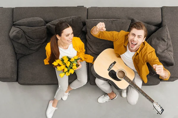 Overhead view of man with guitar raising fists and shouting while his girlfriend holding bouquet on couch at home — Stock Photo