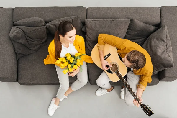 Vue aérienne de l'homme jouant de la guitare à sa petite amie alors qu'elle tient un bouquet sur le canapé à la maison — Photo de stock