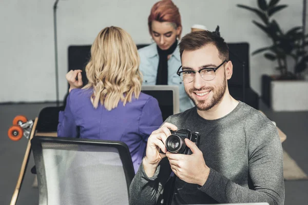 Вибірковий фокус усміхненого фотографа, який дивиться на камеру з колегами позаду в офісі — стокове фото