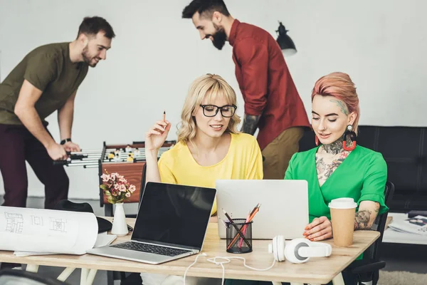 Kreative Geschäftsfrauen arbeiten gemeinsam an einem Projekt, während Kollegen im Büro hintereinander Tischkicker spielen — Stockfoto