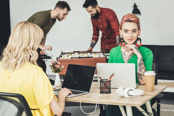 Foco seletivo de jovens designers sentados no local de trabalho com colegas enquanto empresários que jogam futebol de mesa no escritório — Fotografia de Stock