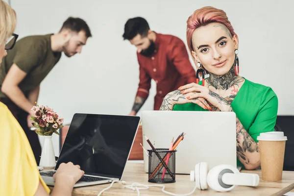 Selective focus of young designer sitting at workplace with colleague while businessmen playing table football in office — Stock Photo