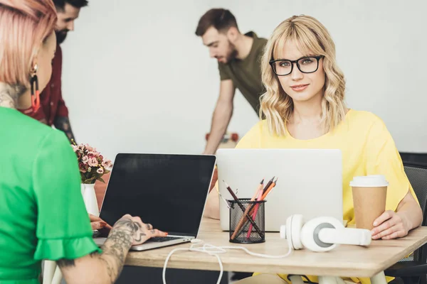 Foco seletivo de jovens blogueiros em óculos sentados no local de trabalho e trabalhadores criativos no escritório — Fotografia de Stock