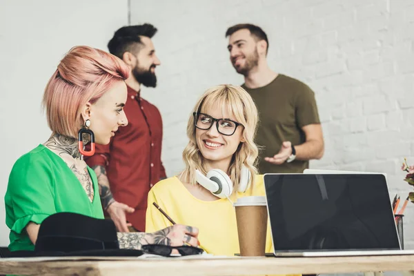 Souriantes femmes d'affaires créatives travaillant ensemble sur le projet tandis que des collègues parlent derrière dans le bureau — Photo de stock