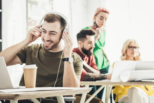 Foco seletivo de homem sorridente em fones de ouvido com café para ir e colegas criativos que trabalham para trás no escritório — Fotografia de Stock