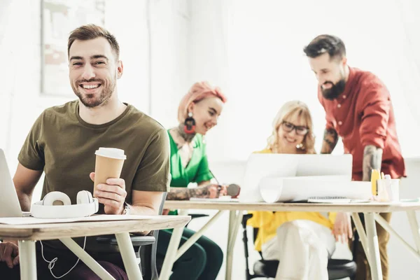 Foyer sélectif de sourire homme avec du café à emporter et des collègues créatifs travaillant derrière dans le bureau — Photo de stock