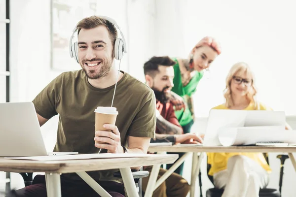 Selektiver Fokus eines lächelnden Mannes mit Kopfhörern, Coffee to go und kreativen Kollegen, die im Büro hinterherarbeiten — Stockfoto