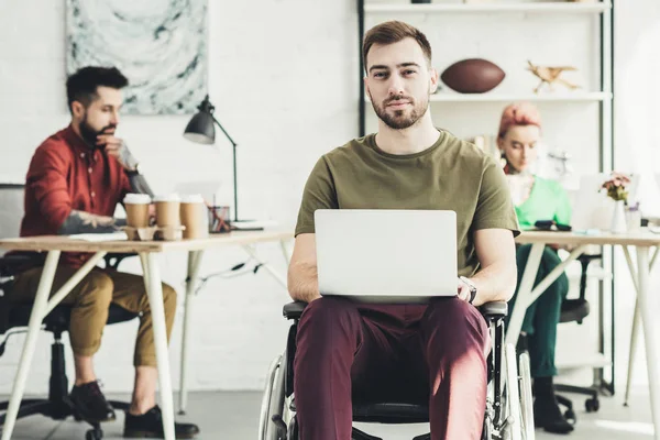 Selective focus of smiling disabled man with laptop and colleagues working behind in office — Stock Photo