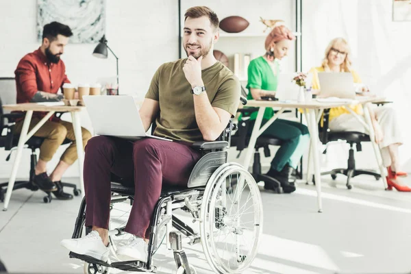 Selective focus of smiling disabled businessman with laptop and colleagues working behind in office — Stock Photo