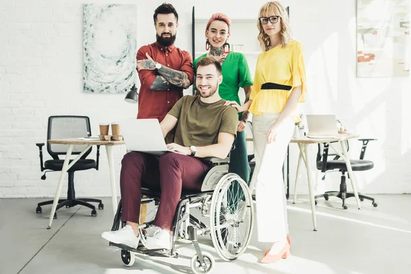 Homme d'affaires handicapé souriant avec ordinateur portable et collègues debout derrière dans le bureau — Photo de stock