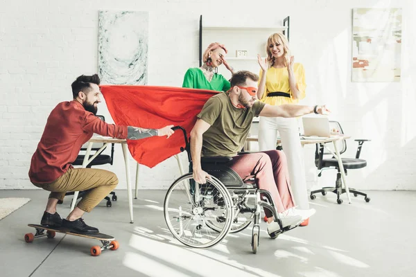 Empresário deficiente em traje de super-herói com colega no skate e empresárias atrás no escritório — Fotografia de Stock