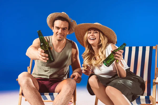 Jeune couple avec des bouteilles de bière assis dans des chaises longues sur fond bleu — Photo de stock