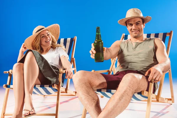 Hombre sosteniendo la botella de cerveza y sentado en la silla de playa sobre fondo azul - foto de stock