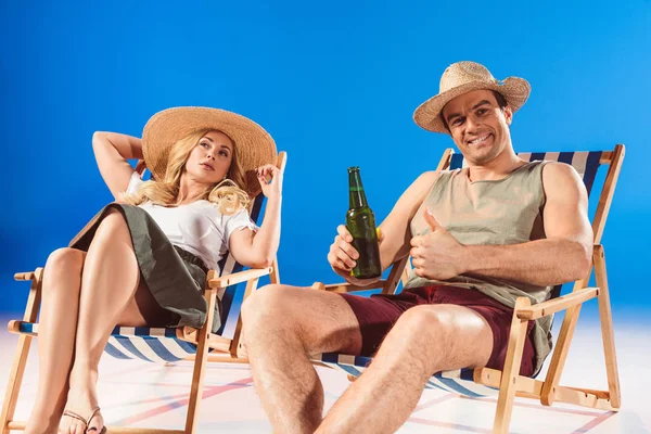 Jovem sorridente com cerveja e mulher relaxando em cadeiras de praia em fundo azul — Fotografia de Stock