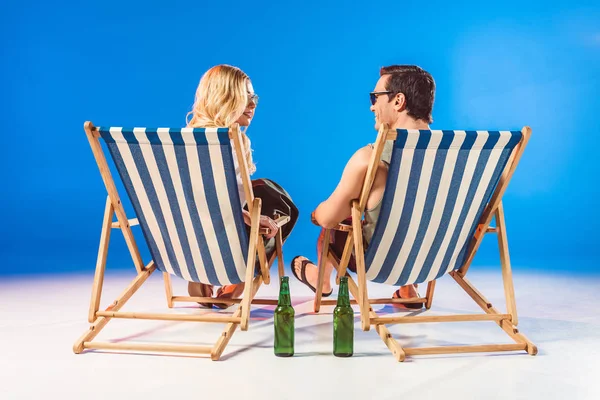Усміхнена молода пара розслабляється в стільцях палуби пляшками пива на синьому фоні — стокове фото