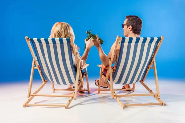 Couple souriant en lunettes de soleil toastant avec des bouteilles de bière reposant sur des chaises longues sur fond bleu — Photo de stock