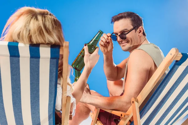 Homme en lunettes de soleil regardant la bouteille de bière à la main féminine alors qu'il était assis sur une chaise de plage isolé sur bleu — Photo de stock