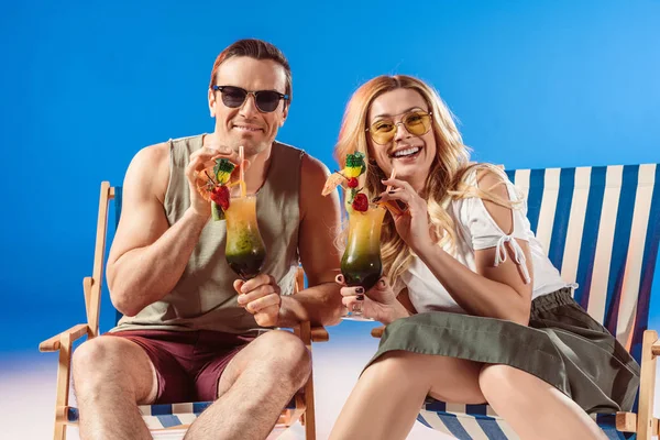 Homem e mulher bebendo coquetéis tropicais descansando em cadeiras de praia em fundo azul — Fotografia de Stock