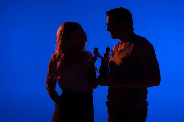 Silhouette of beautiful woman and man holding cocktails isolated on blue in dark light — Stock Photo