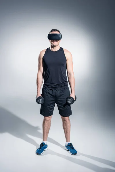 Hombre deportivo haciendo ejercicio con pesas mientras usa anteojos vr sobre fondo gris - foto de stock
