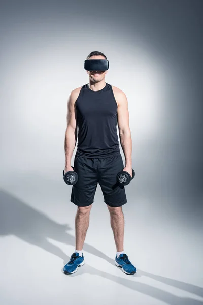 Sportive man exercising with dumbbells while wearing vr glasses on grey background — Stock Photo