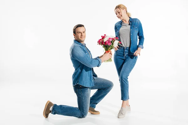 Petit ami debout sur un genou et donnant femme blonde un bouquet de fleurs isolées sur blanc — Photo de stock